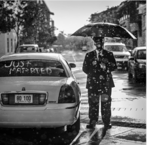 A stylish man, umbrella in hand, stands beside a parked car.